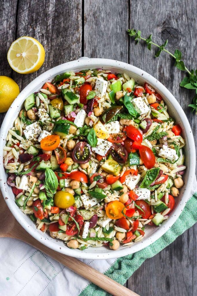 Greek orzo pasta salad in large bowl with cubed feta, sliced cherry tomatoes, and fresh basil.