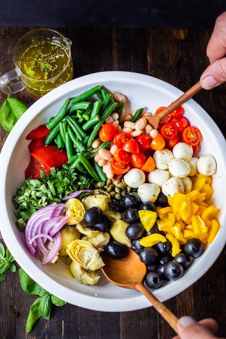 Antipasto Salad wtih white beans, aritchoke hearts, olives, fresh veggies, and lots of herbs in large salad bowl with serving utensils.