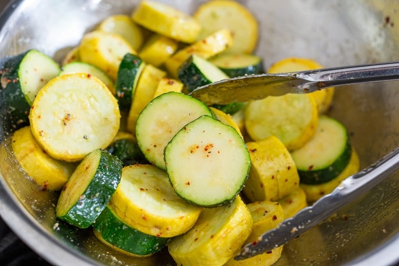 zucchini and yellow squash in mixing bowl with tongs, tossed with olive oil, salt, pepper, and chili flakes.