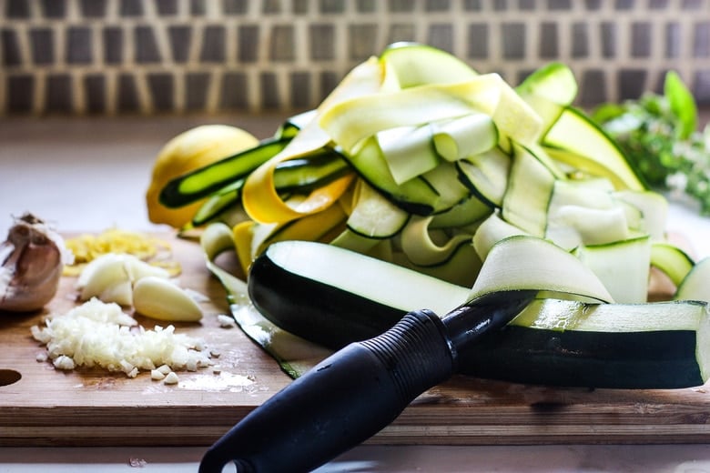 using vegetable peeler on zucchini next to pile of zucchini ribbons on board with minced garlic.