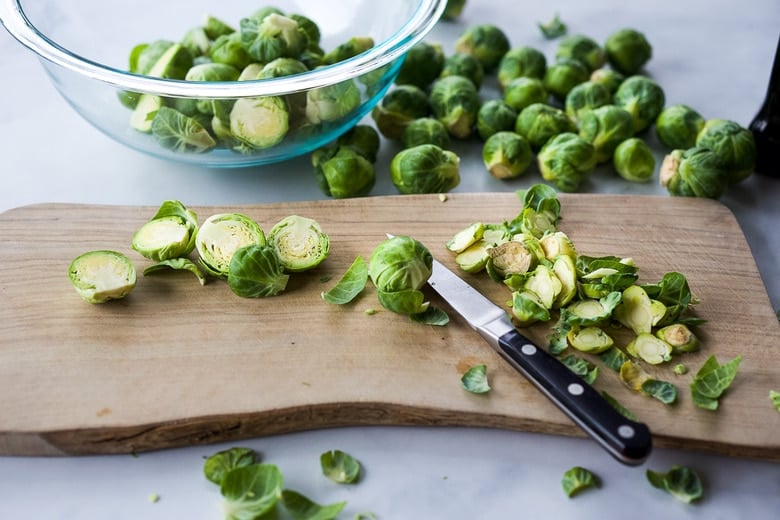 trimming and slicing Brussels sprouts in half on a wood cutting board with a knife before tossing in a glass bowl.