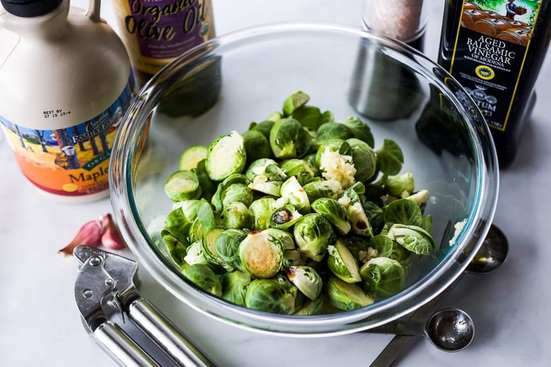 tossing brussels sprouts with seasonings in a glass mixing bowl, surrounded by spoons, garlic press, balsamic, salt, olive oil, and maple syrup.