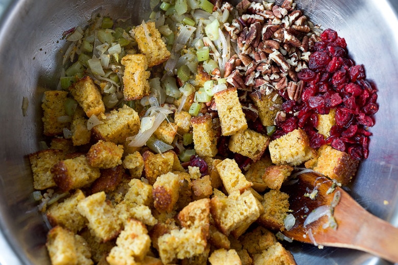 mixing together ingredients for cornbread stuffing in a mixing bowl - cubed cornbread, cranberries, pecans, and sautéed vegetables.