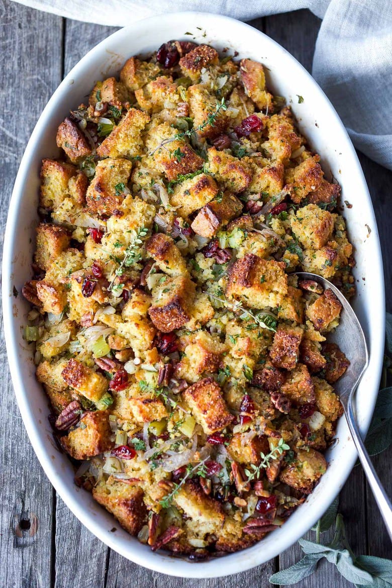 oval baking dish with cornbread stuffing with serving spoon, garnished with fresh herbs.