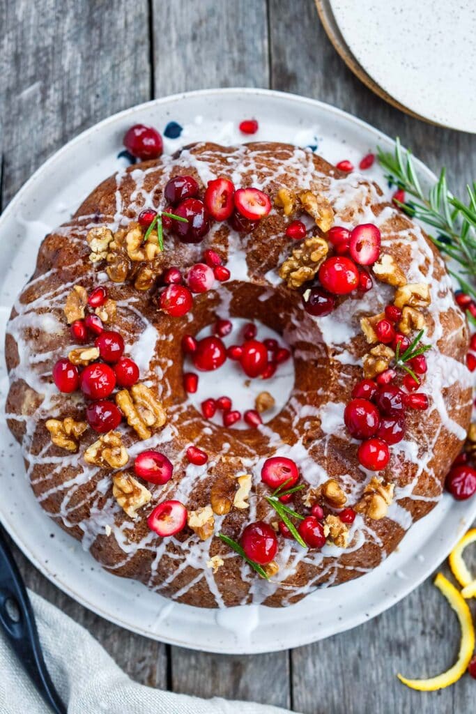 Vegan Cranberry Cake in a bundt shape, drizzled with a sugar glaze and topped with fresh cranberries, pomegranates, walnuts, and rosemary.