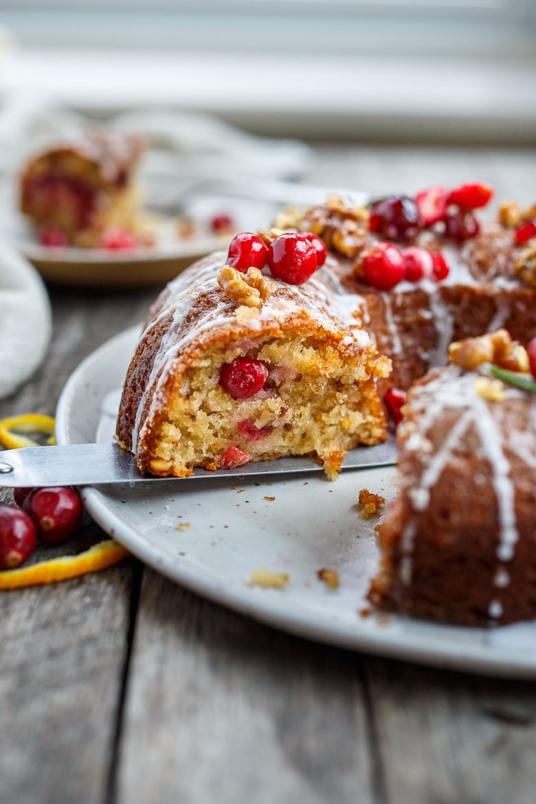 metal spatula removing a slice of vegan cranberry cake with fresh cranberries and powdered sugar glaze.