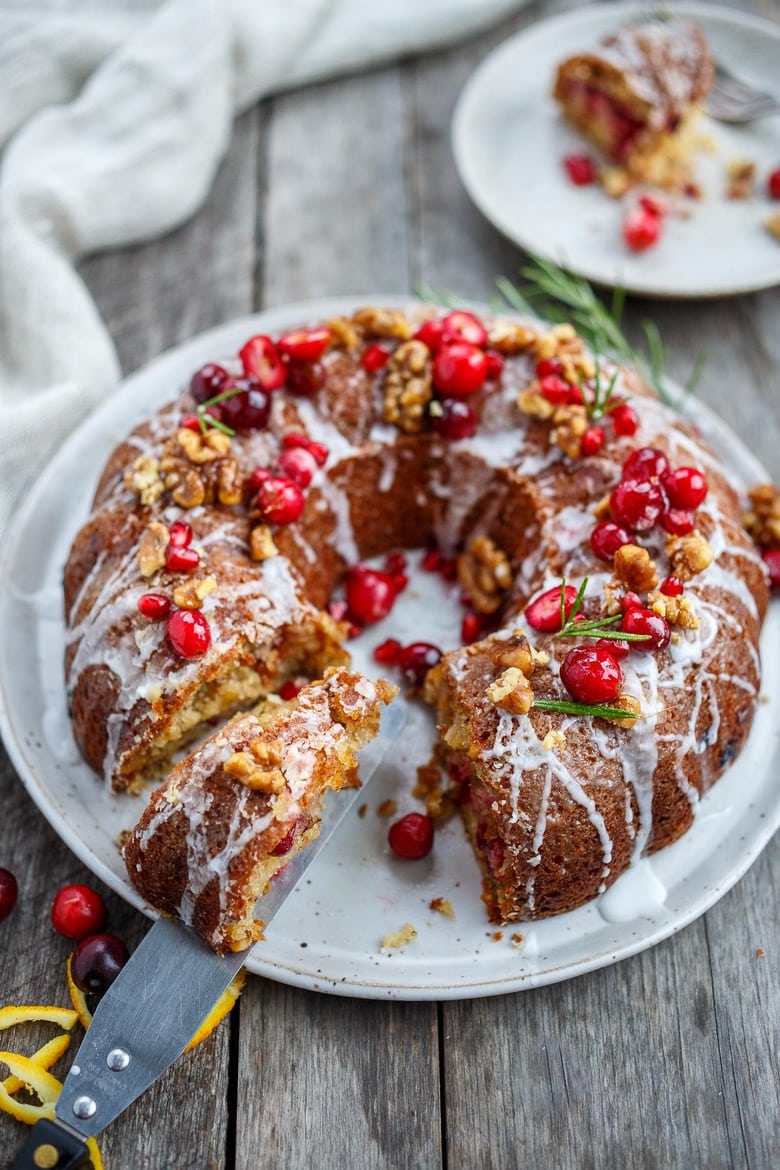 vegan cranberry cake in bundt shape on plate decorated with drizzle of glaze, fresh cranberries, walnuts, and rosemary with a spatula removing a slice.