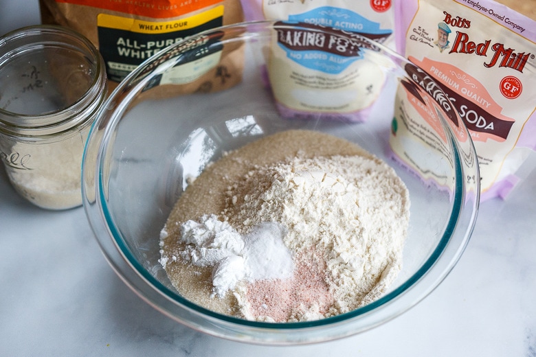 mixing dry ingredients together in a glass bowl next to ingredients in their containers - baking soda, baking powder, all-purpose white wheat flour, sugar.