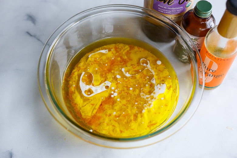 mixing liquid ingredients together in a glass bowl next to orange juice, vanilla extract, and olive oil.