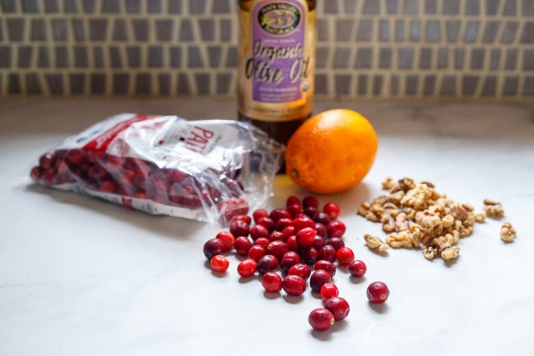 some ingredients for cranberry cake laid on kitchen counter - olive oil, cranberries, orange, and walnuts.