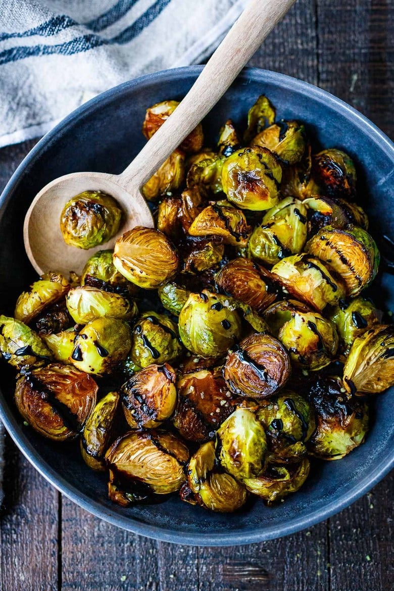 serving bowl with roasted brussels sprouts and a drizzle of balsamic glaze over top, with a wood serving spoon.