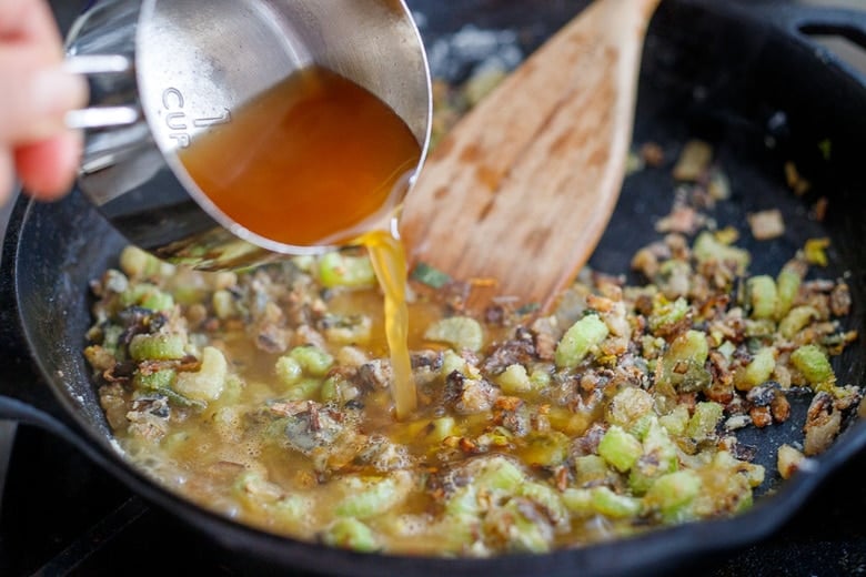 pouring cup of vegetable broth into cast iron skillet with sautéed celery and onion.
