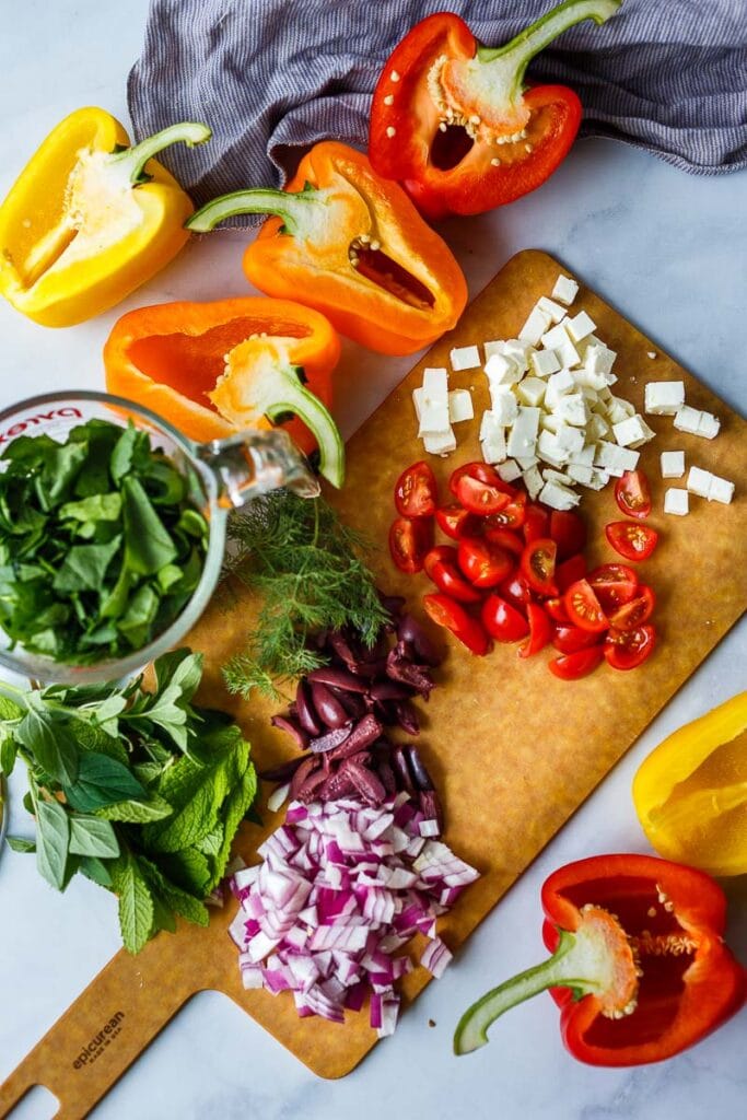 chopped ingredients for mediterranean peppers on cutting board- feta, tomatoes, olives, onion, herbs.