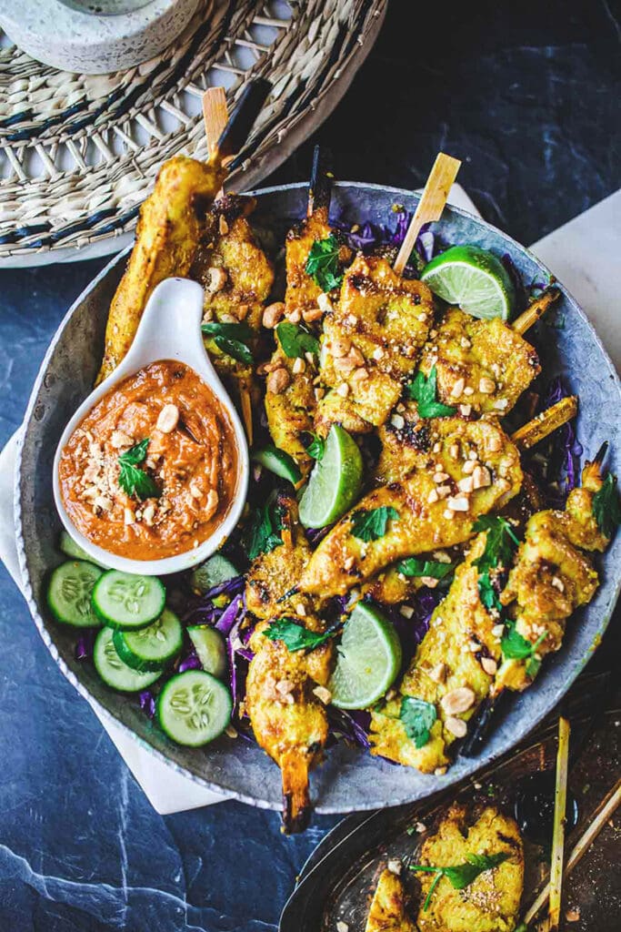 grilled chicken satay on skewers in bowl with sliced purple cabbage and cucumber, along with lime wedges, crushed peanuts, and a small dish of peanut sauce.