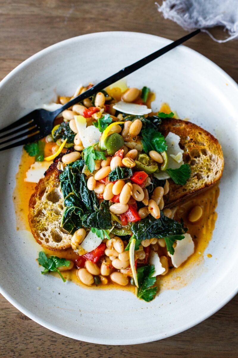 Brothy Beans with lacinato kale, roasted peppers, olives, capers and lemon zest over garlic-infused Sourdough Bread -a simple pantry meal that can be made in under 30 minutes! Vegan adaptable.