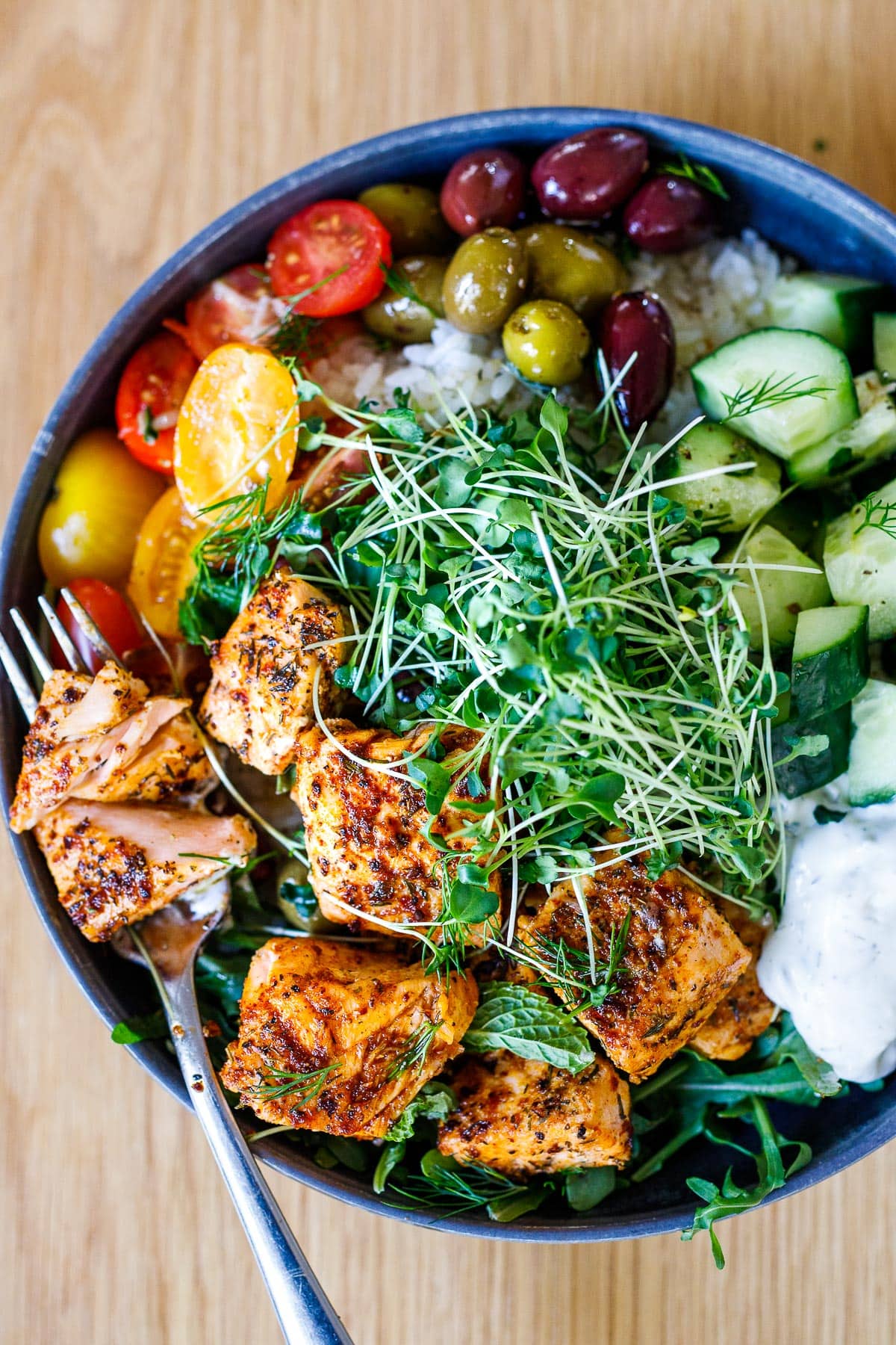 Delicious and healthy Greek Salmon Bowl with rice, greens, olives, tomatoes, cucumber, tzatziki and greek dressing.