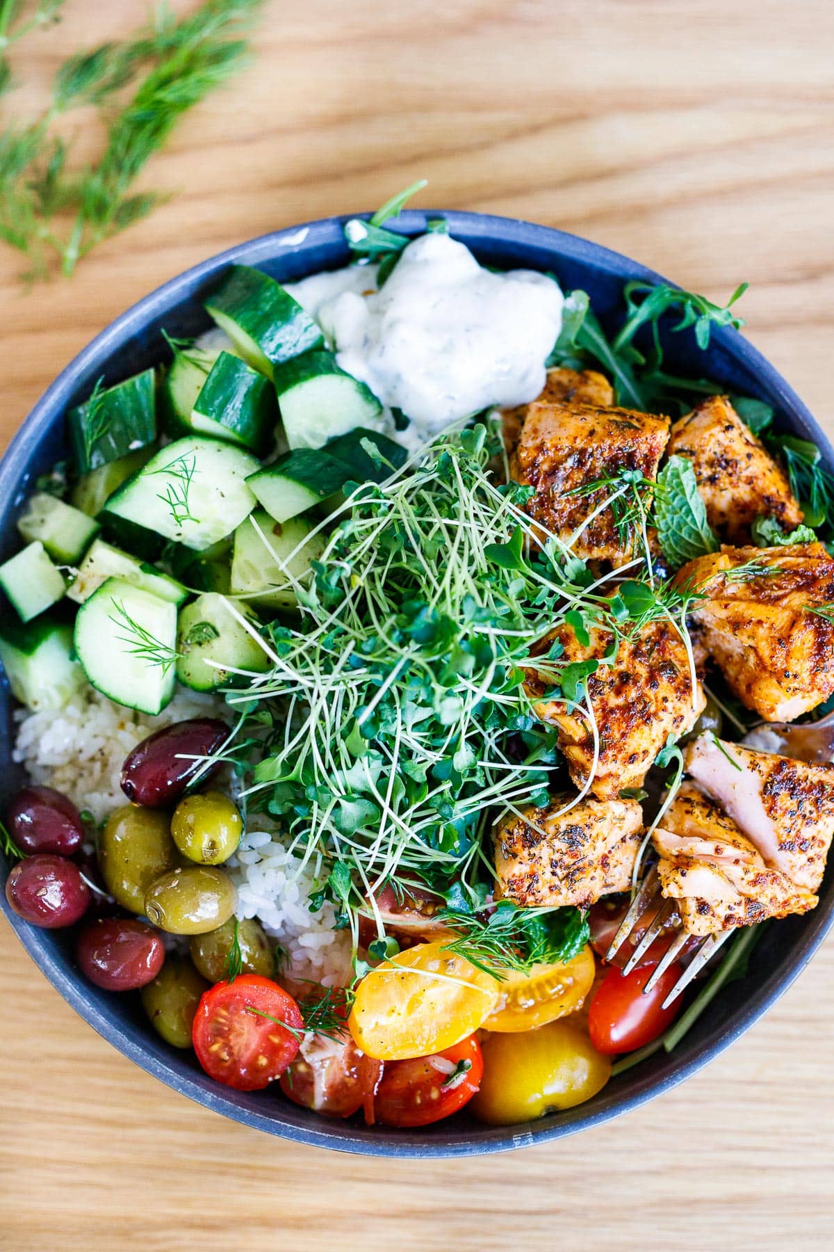 A Greek Salmon Bowl with rice, greens, olives, tomatoes, cucumber, tzatziki and greek dressing.