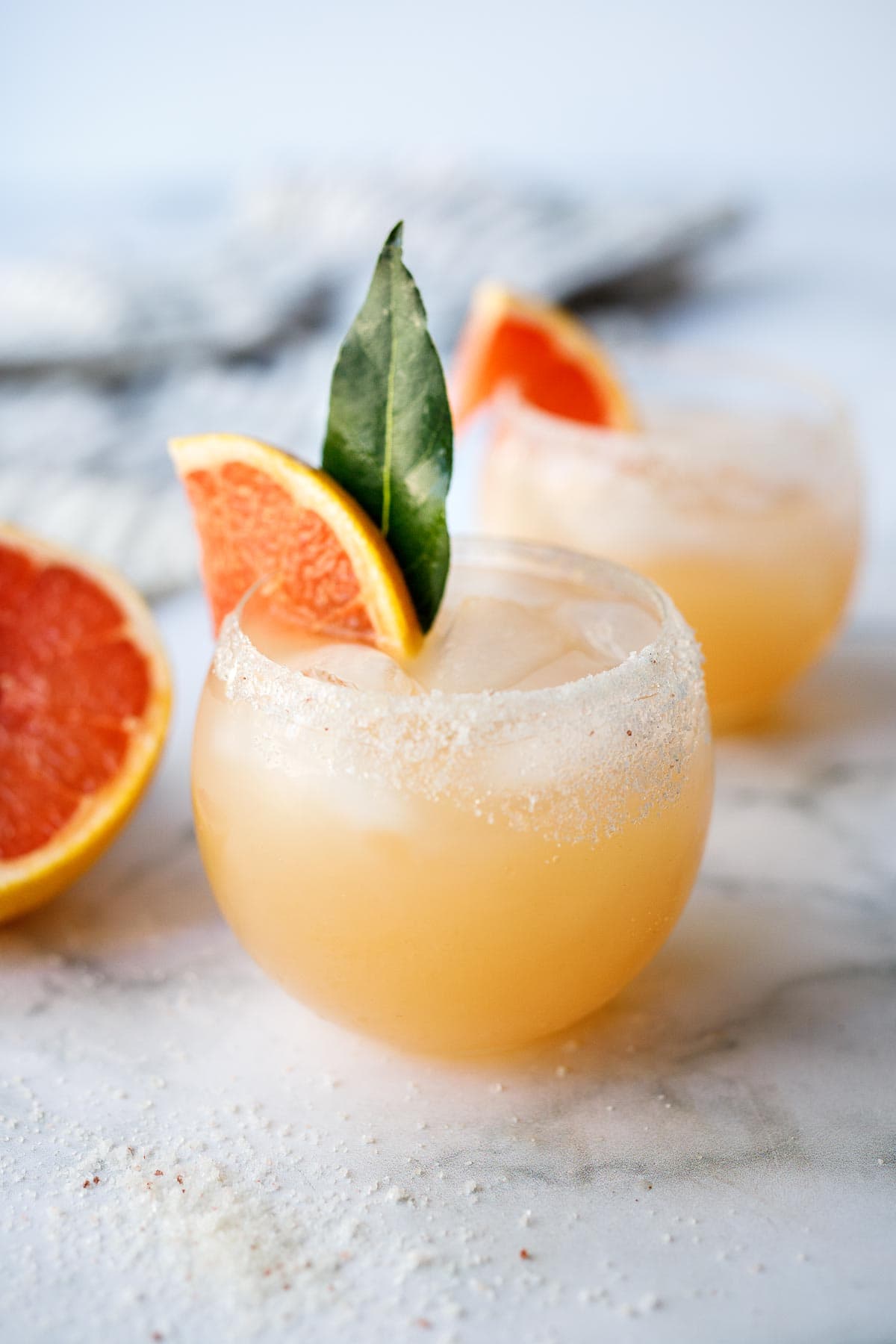 two glasses of paloma cocktails with salted rims, garnished with grapefruit wedge and bay leaf.