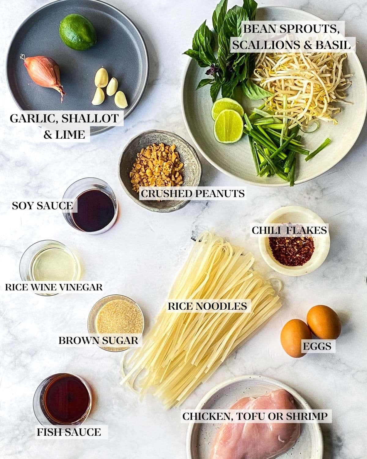 Pad thai ingredients  on a white marble counter;  rice noodles, garlic, shallots, limes, bean sprouts, thai basil, crushed peanuts, chili flakes, brown sugar, rice vinegar, soy sauce, eggs and fish sauce. 