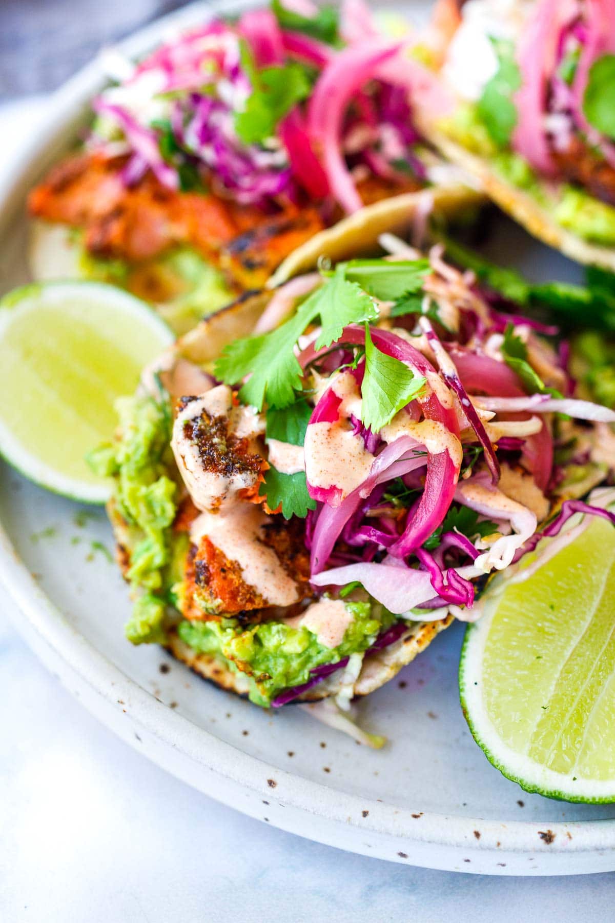salmon tacos with mashed avocado and pickled onions and cabbage-fennel slaw, topped with cilantro, served on plate with lime halves.