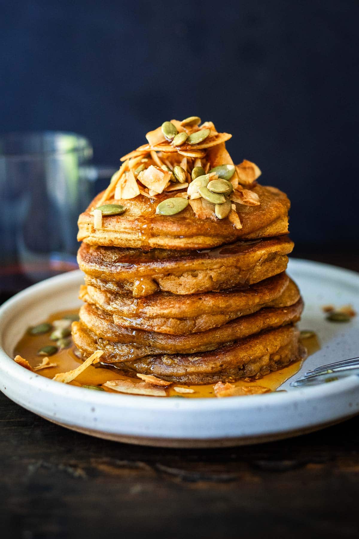 pumpkin pancakes stacked on a white place with maple syrup, coconut flakes and pumpkin seeds.