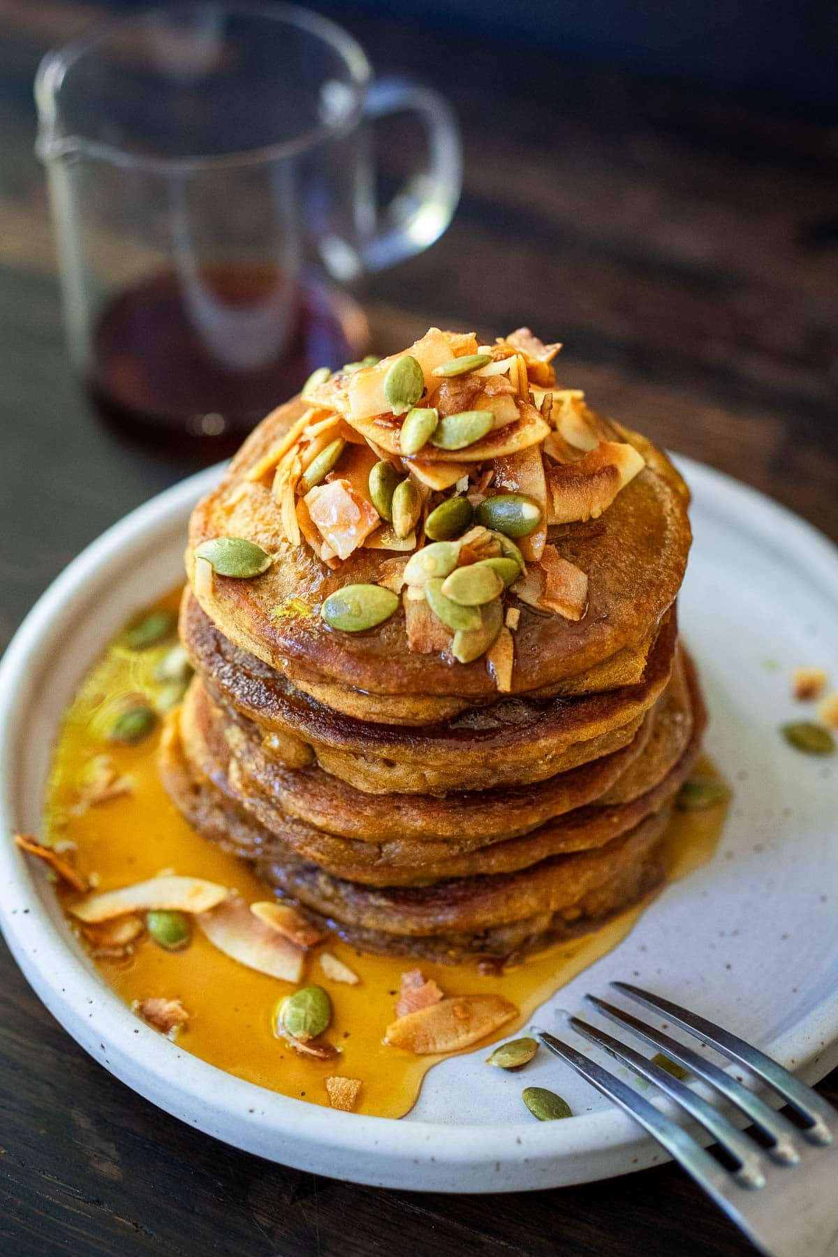 pumpkin pancakes stacked on a white place with maple syrup, coconut flakes and pumpkin seeds.