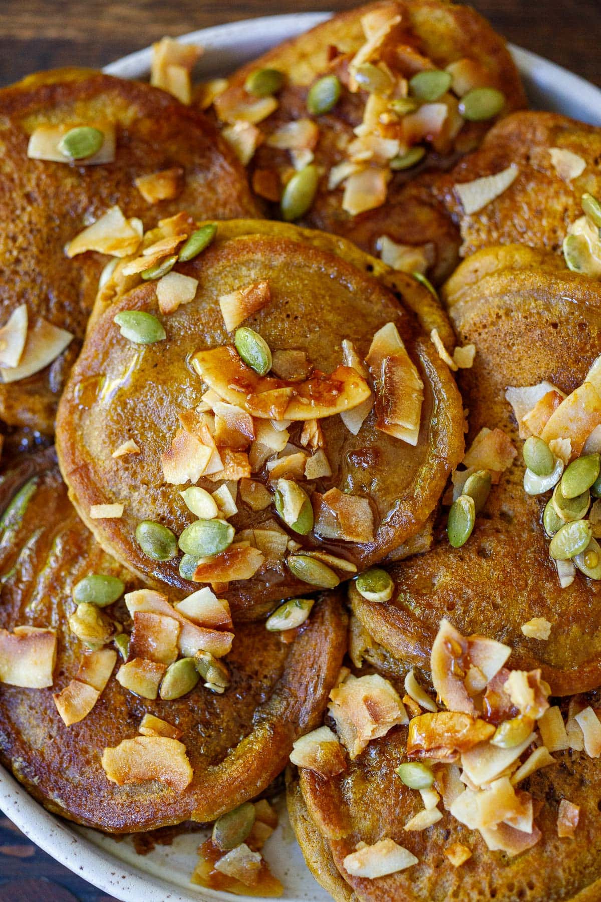 pumpkin pancakes stacked on a white place with maple syrup, coconut flakes and pumpkin seeds.