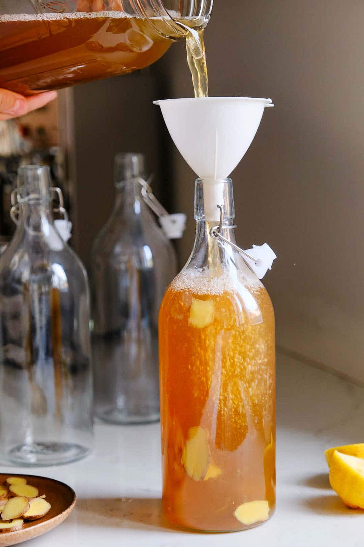 pouring kombucha through funnel into glass bottle with sliced ginger.