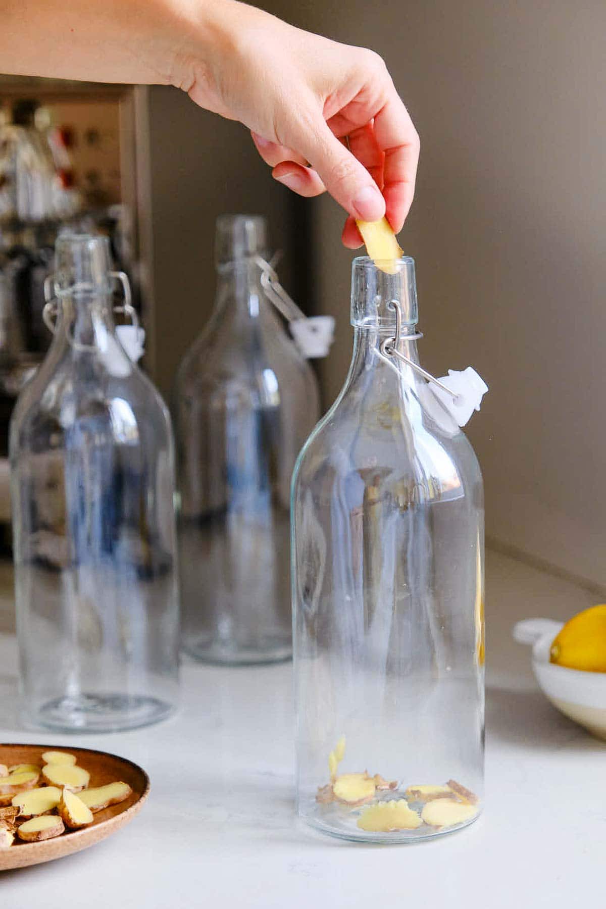 glass bottles on countertop - hand adding sliced ginger into one bottle.