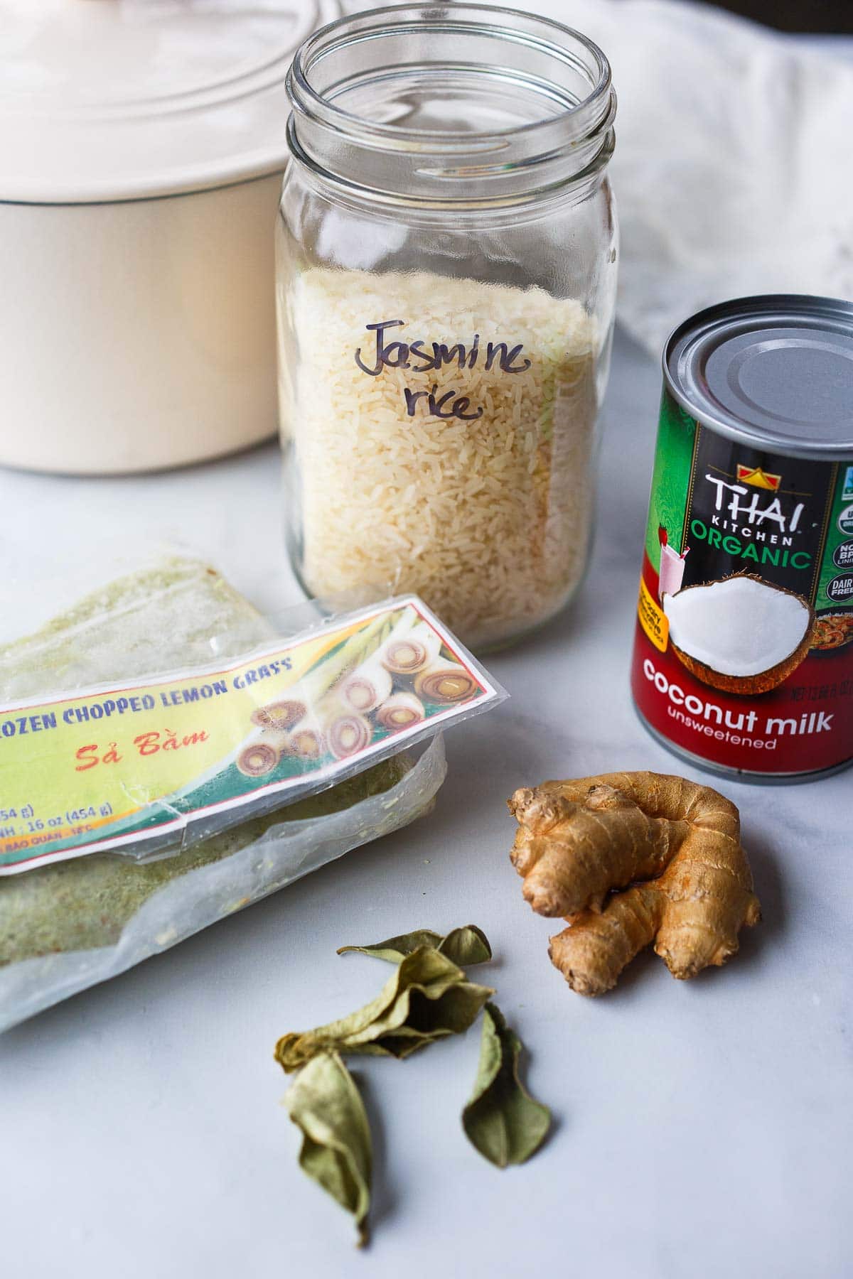 ingredients laid out for coconut rice- jar of jasmine rice, can of coconut milk, bag of frozen chopped lemon grass, fresh ginger, and dried lime leaves.