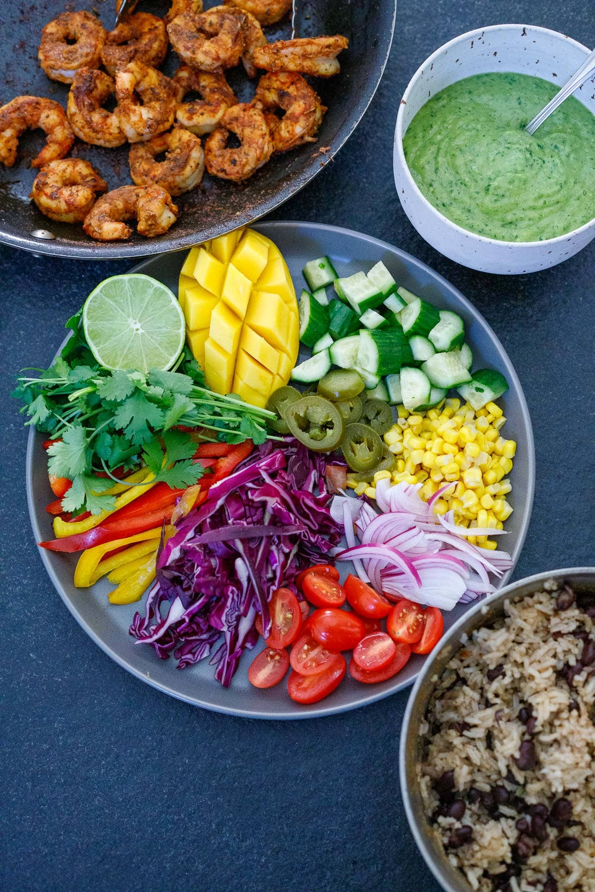 ingredients for spicy mexican shrimp and rice bowls - skillet with shrimp, creamy avocado sauce, plate with chopped and sliced veggies, mango, and herbs, and pot with rice and beans.