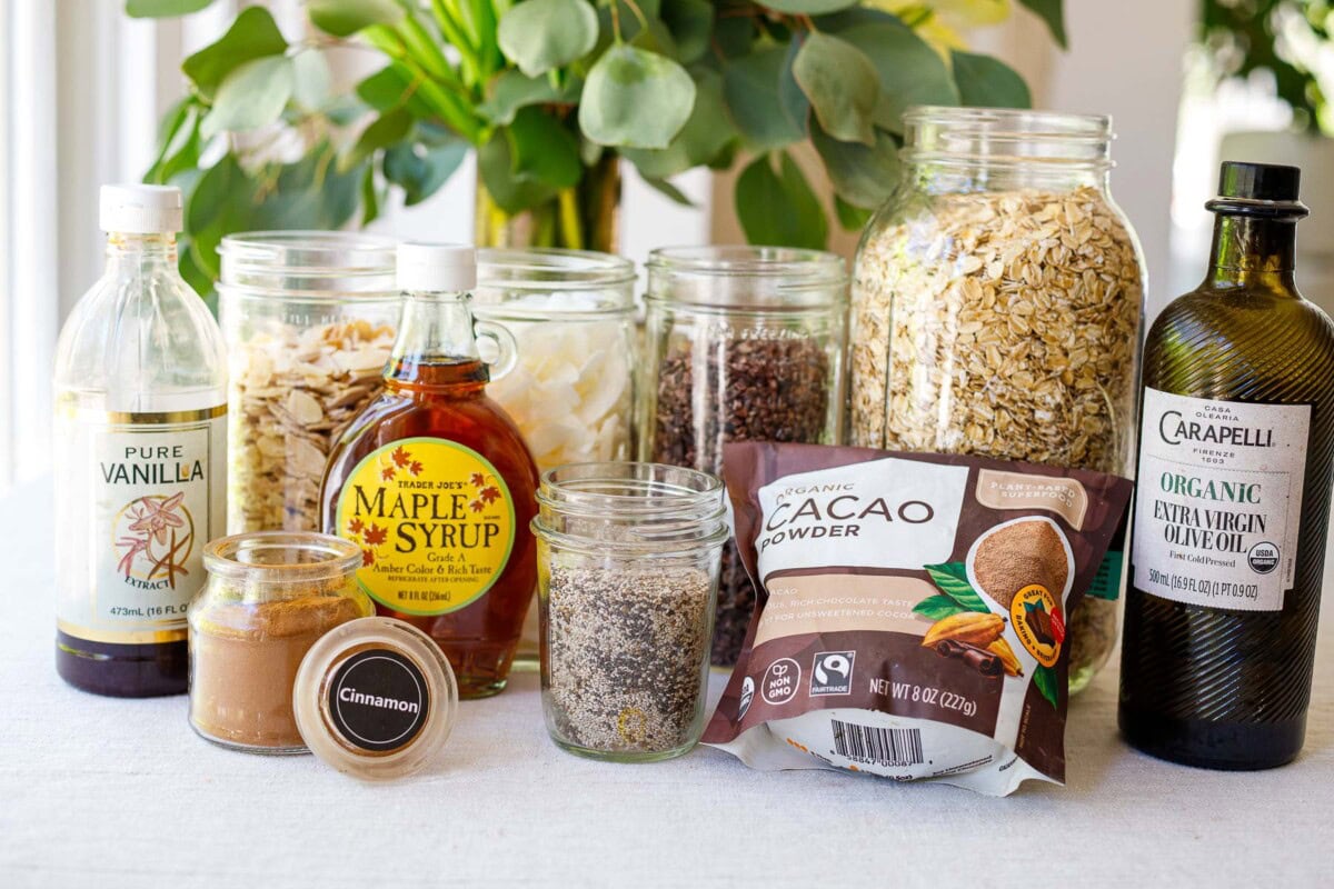 ingredients laid out for dark chocolate granola- mason jars with oats, cacao nibs, coconut flakes, flax and chia seeds, vanilla extract, cinnamon, maple syrup, cacao powder, and olive oil.