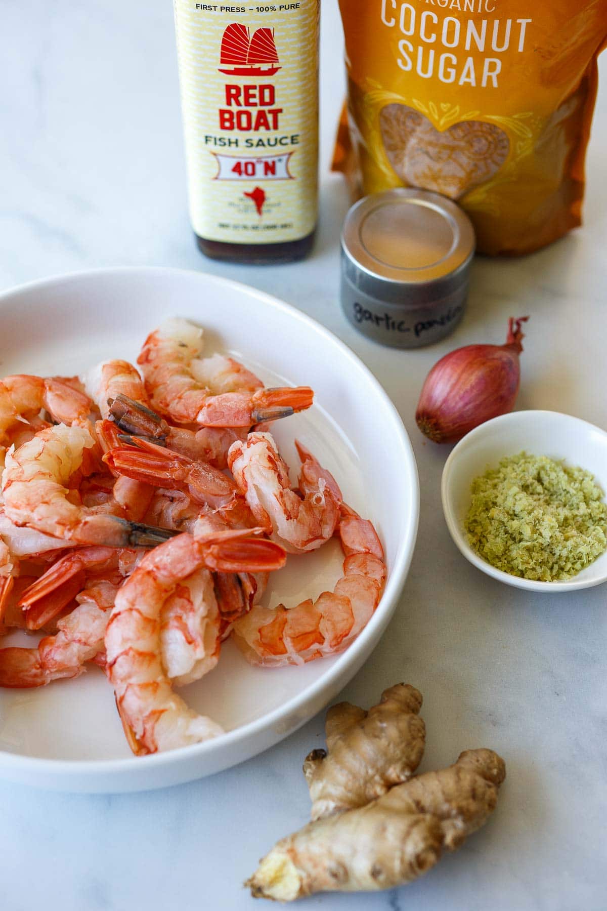 bowl of raw shrimp next to ingredients like fresh ginger, lemongrass, shallot, garlic powder, fish sauce, and coconut sugar. 