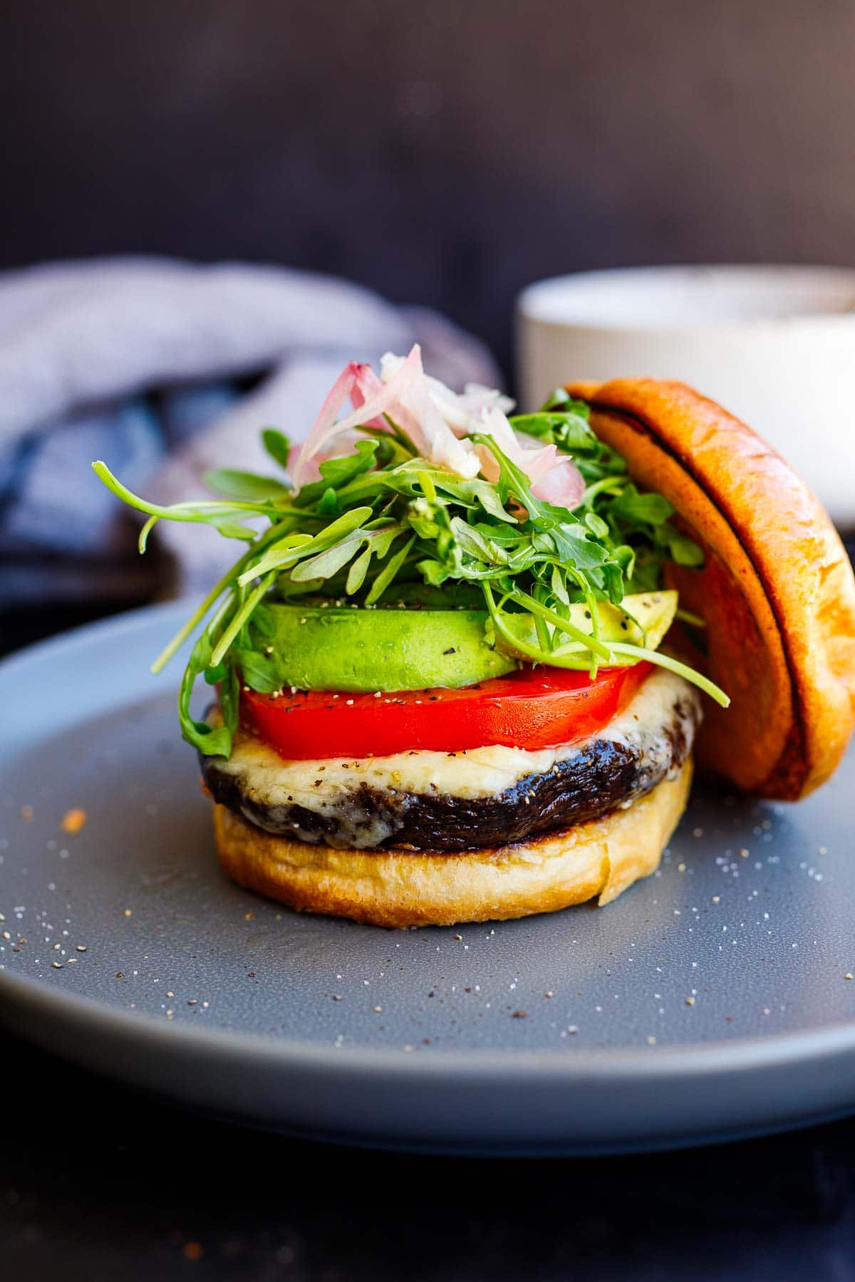 portobello mushroom burger on plate on brioche bun, stacked with melted cheese, sliced tomato and avocado, arugula, and pickled onions.