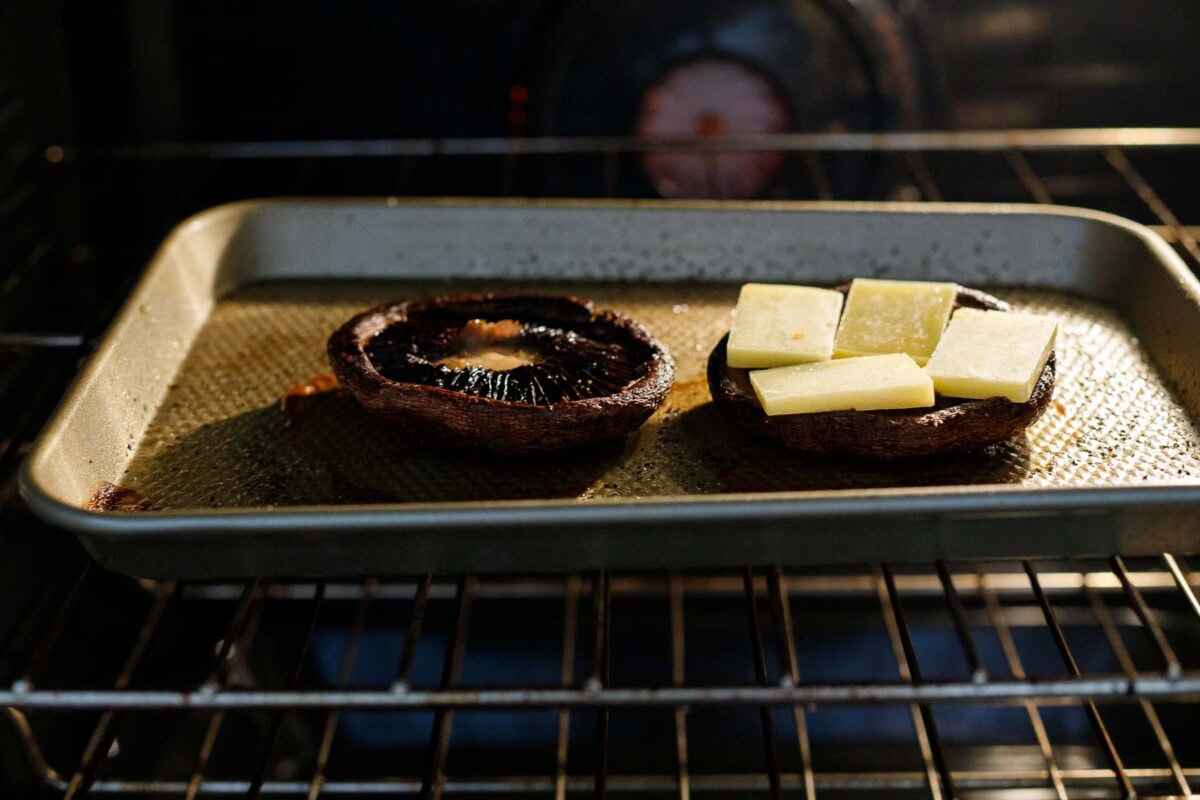 two portobello mushrooms on baking sheet in oven - one with squares of cheese on top.