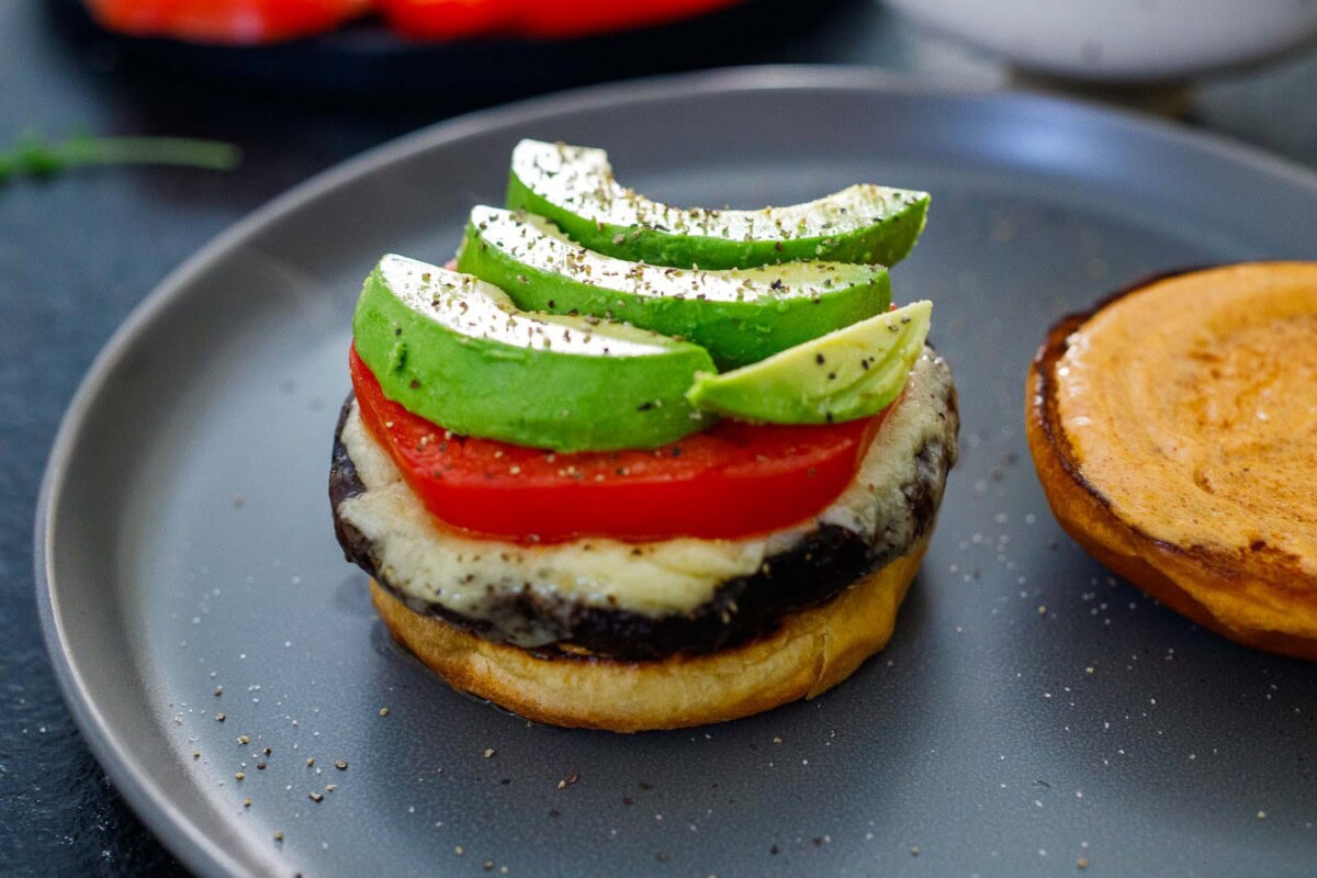 assembling portobello mushroom burger with brioche bun, stacked with roasted portobello, melted cheese, sliced tomato and avocado.