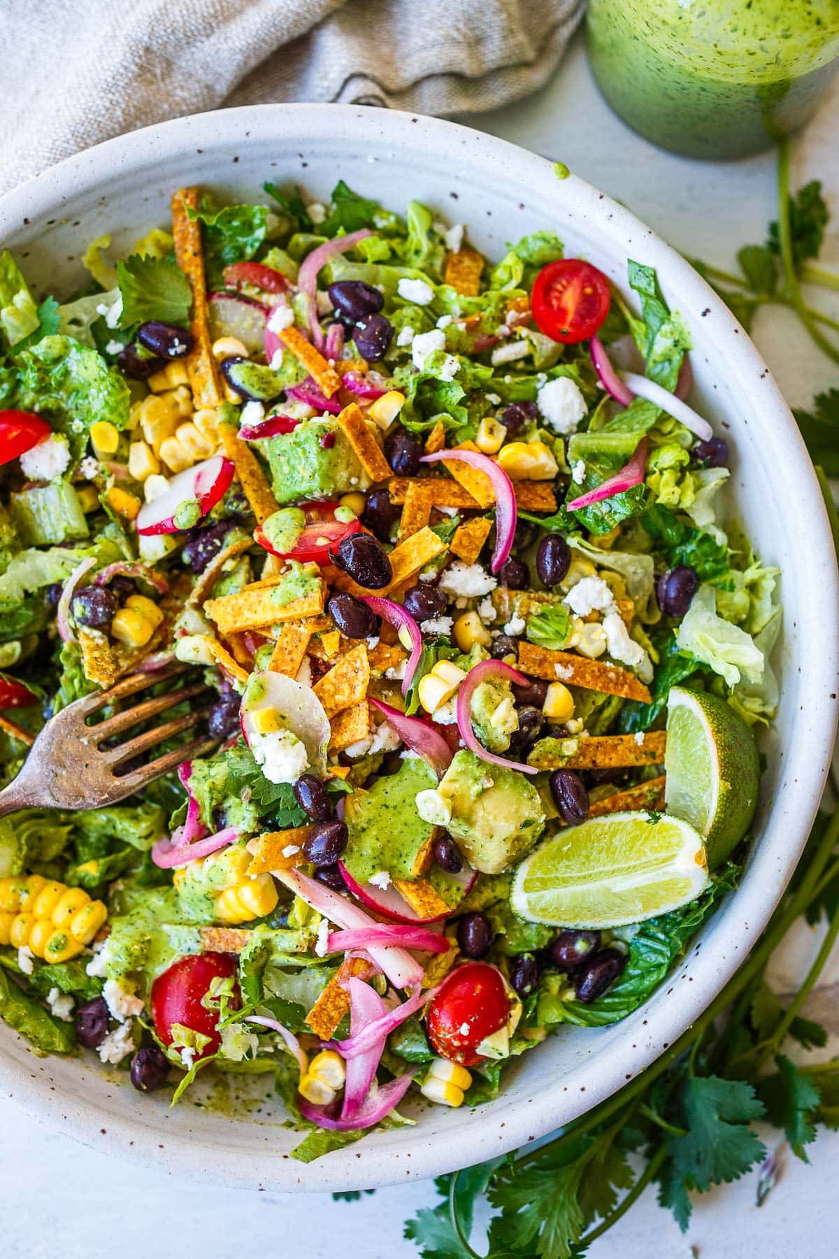 bowl of taco salad with tortilla strips, black beans, pickled onions, radish, lime wedges, corn, cotija cheese, and creamy cilantro lime dressing.