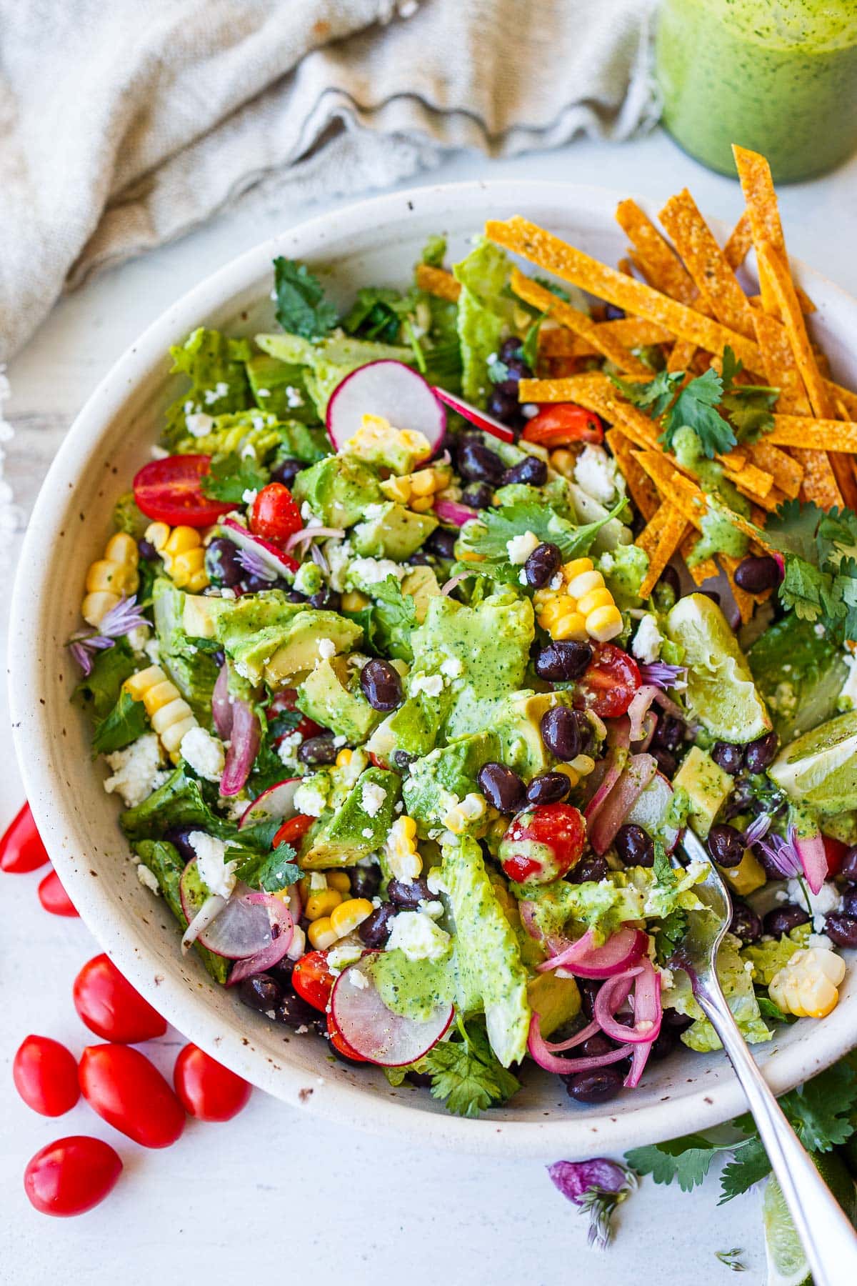 taco salad topped with black beans, corn, cherry tomatoes, avocado, lime wedges, cotija cheese, pickled red onions, radish, and homemade tortilla strips, drizzled in creamy cilantro lime dressing.