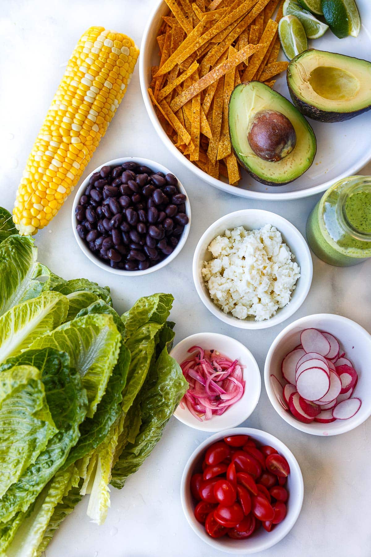 ingredients for taco salad laid out in bowls - tortilla strips, lime wedges, avocado halves, creamy cilantro lime dressing, black beans, cotija cheese, corn on the cob, sliced radish, pickled onions, sliced cherry tomatoes, romaine lettuce.