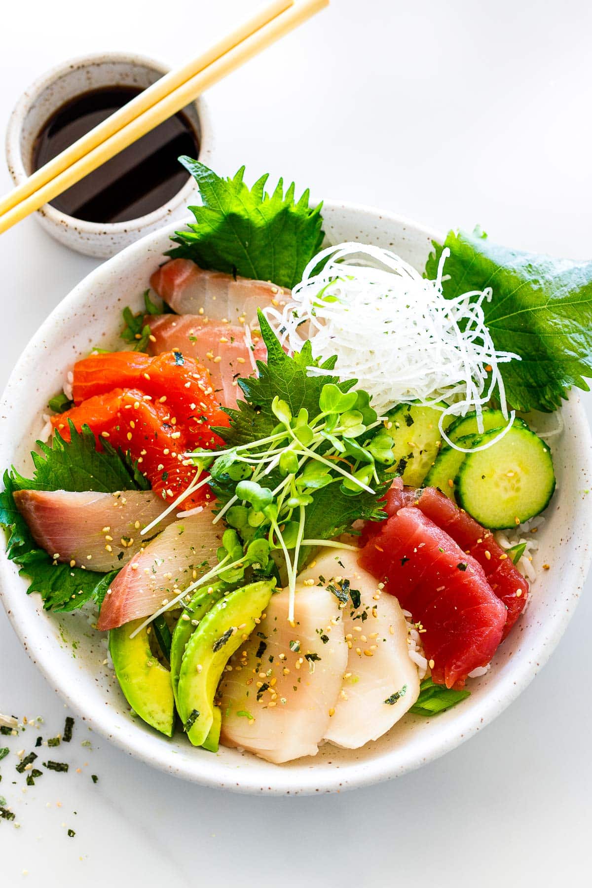 chirashi sushi with sliced fish, sliced cucumber and avocado, grated daikon, shiso leaves, and sprouts, garnished with furikake and toasted sesame seeds.