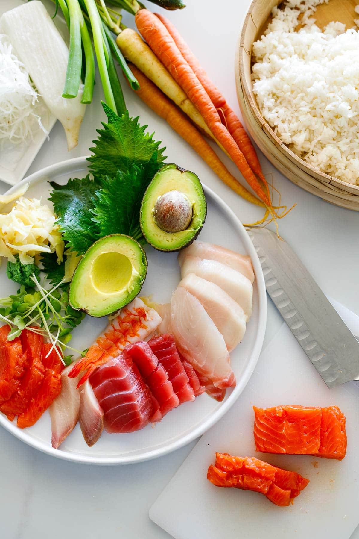 ingredients for chirashi sushi - plate of sliced fish, halved avocado, sprouts, wasabi, pickled ginger, and herbs next to carrot bunch and cooked sushi rice.