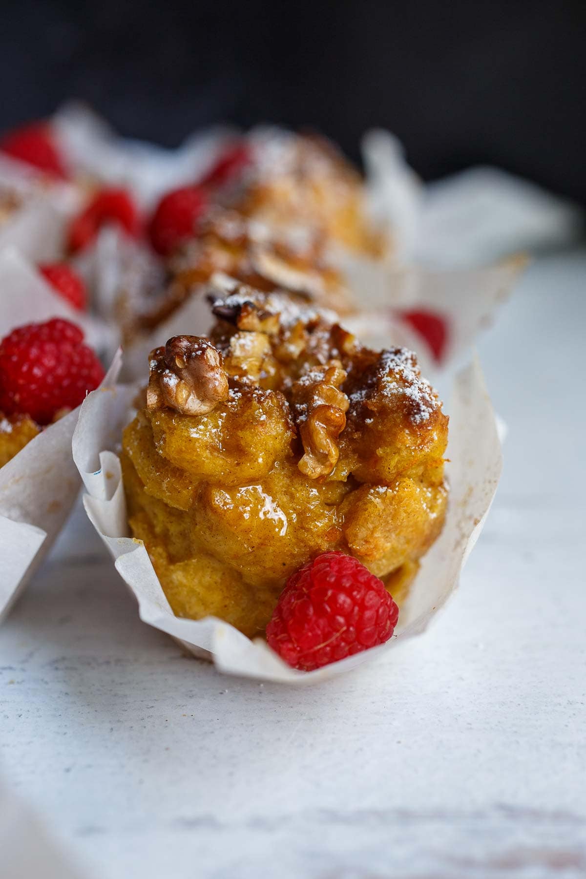 close up of French toast muffin with paper liner opened up to see tender texture, topped with maple syrup, powdered sugar, and fresh raspberries.