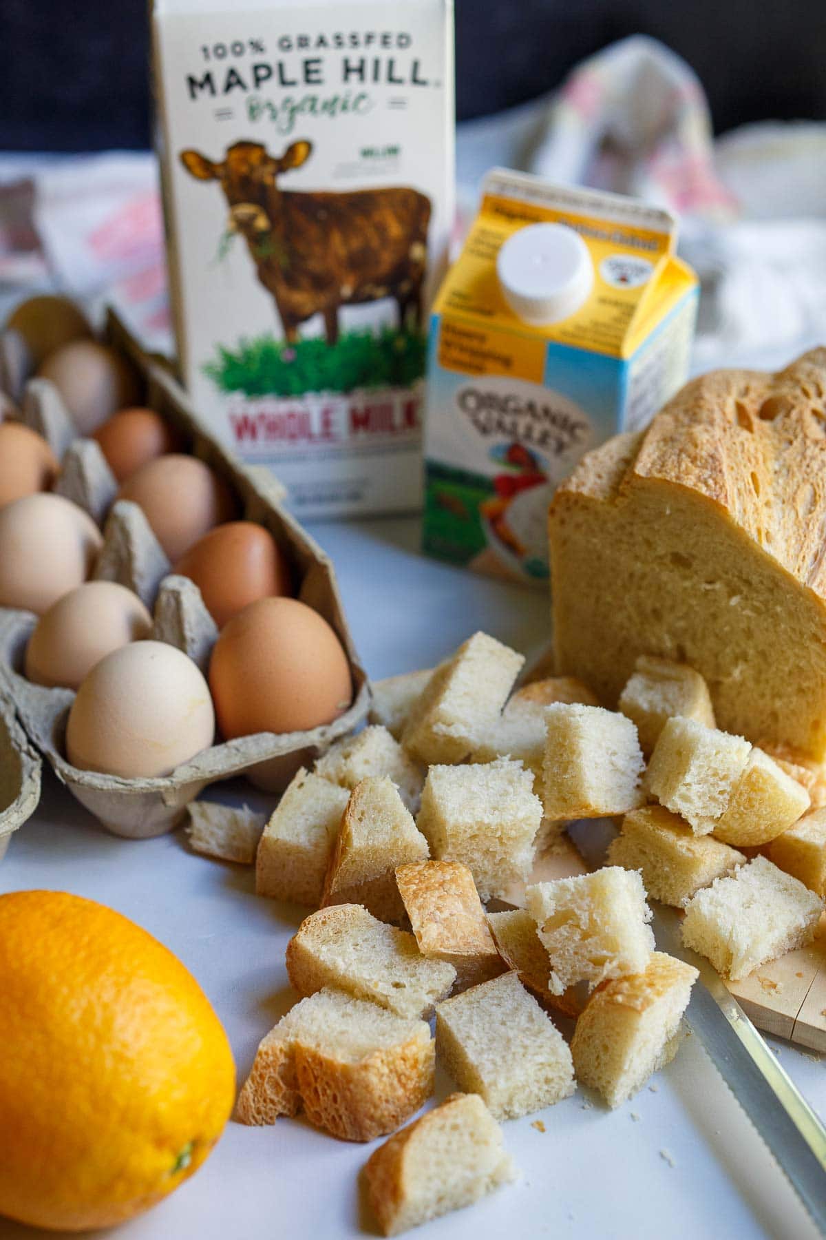 ingredients for French toast muffins laid out - cubed sourdough bread, heavy cream, whole milk, egg carton, and orange.