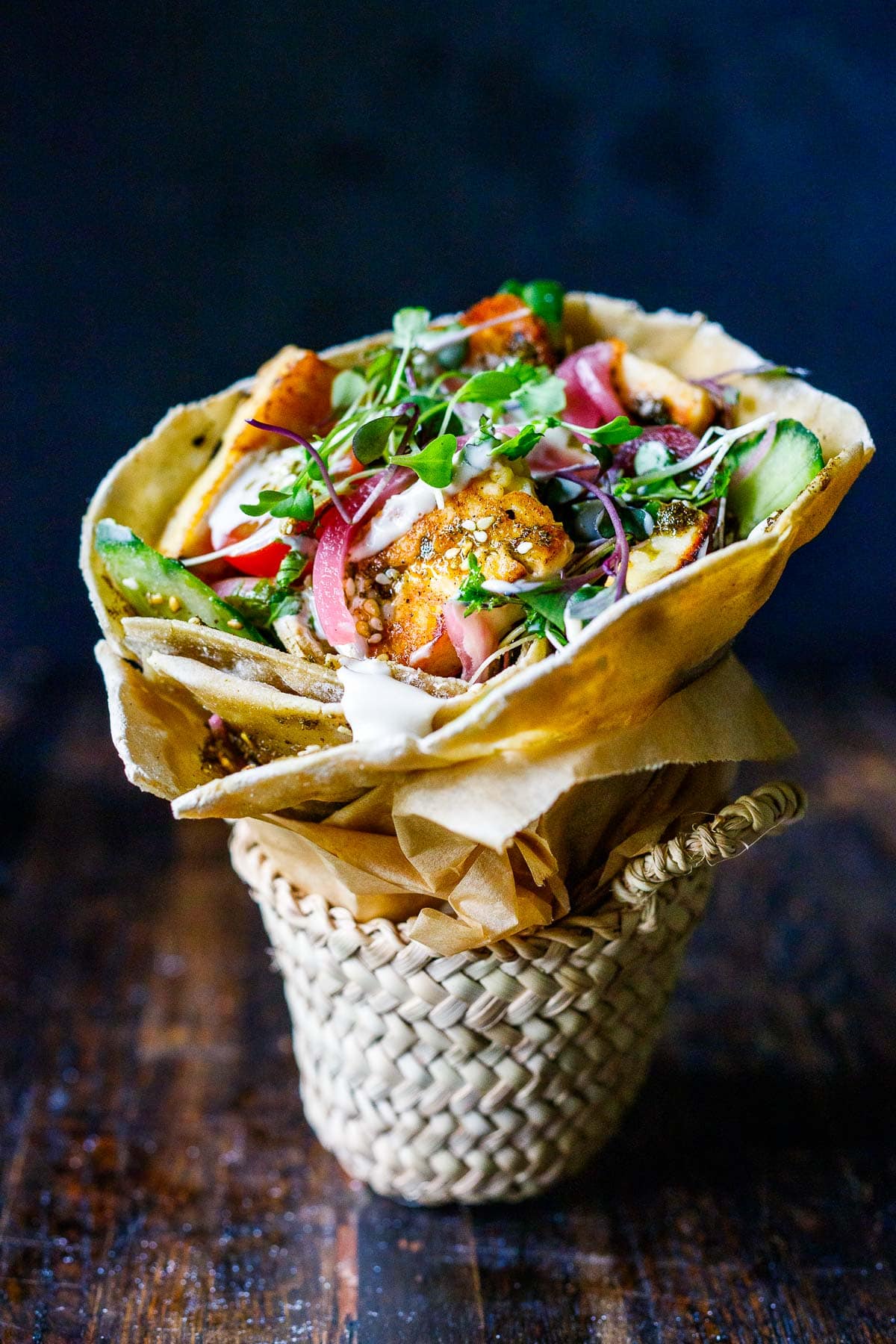 A halloumi wrap with tomatoes, microgreens, cucumbers and tahini yogurt dressing, wrapped in parchment in little basket.