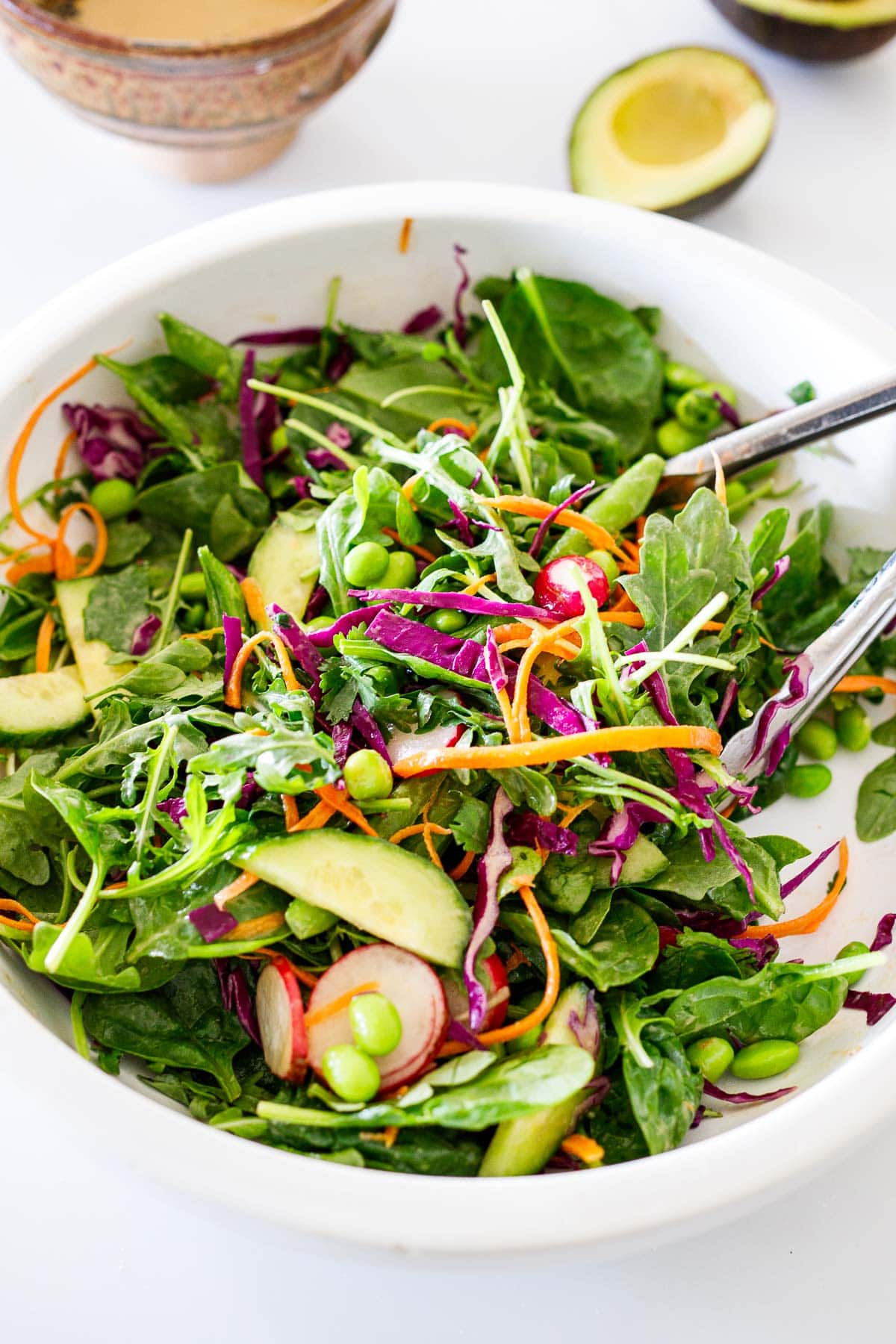 mixing bowl and tongs tossing salad with grated carrots and cabbage, edamame, radish, and cucumber. 