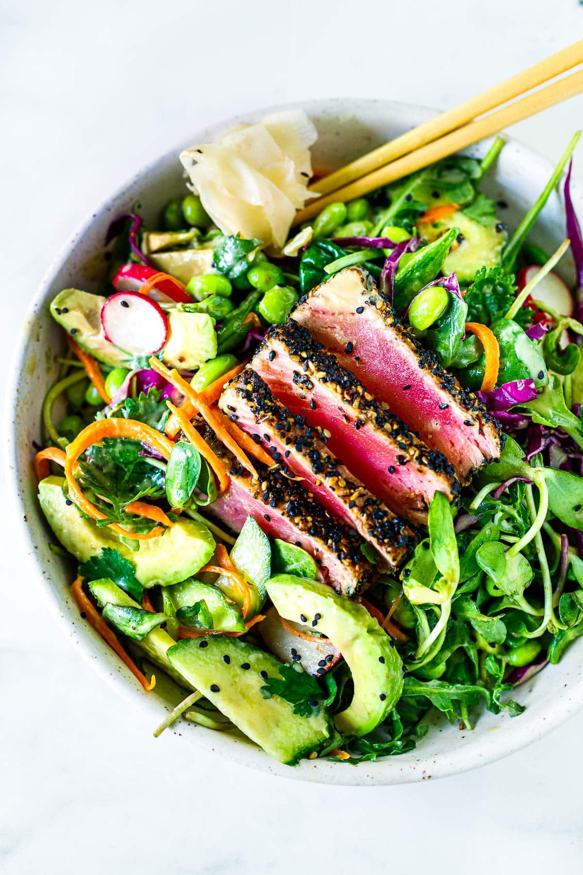 sesame crusted seared ahi tuna salad with sliced avocado, grated carrots, sliced radish, and fresh greens in bowl with pickled ginger and chopsticks. 