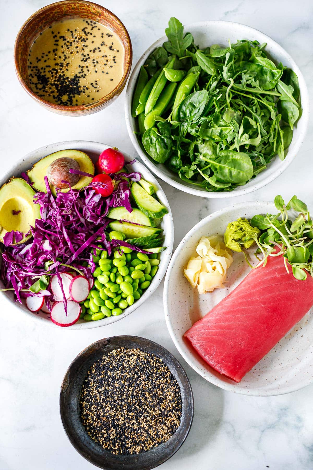 ingredients for seared ahi tuna salad- ginger wasabi dressing, fresh greens, bowl of cabbage, avocado, radish, cucumber, edamame, plate of raw ahi tuna, microgreens, wasabi, pickled ginger, and bowl of white and black sesame seeds. 