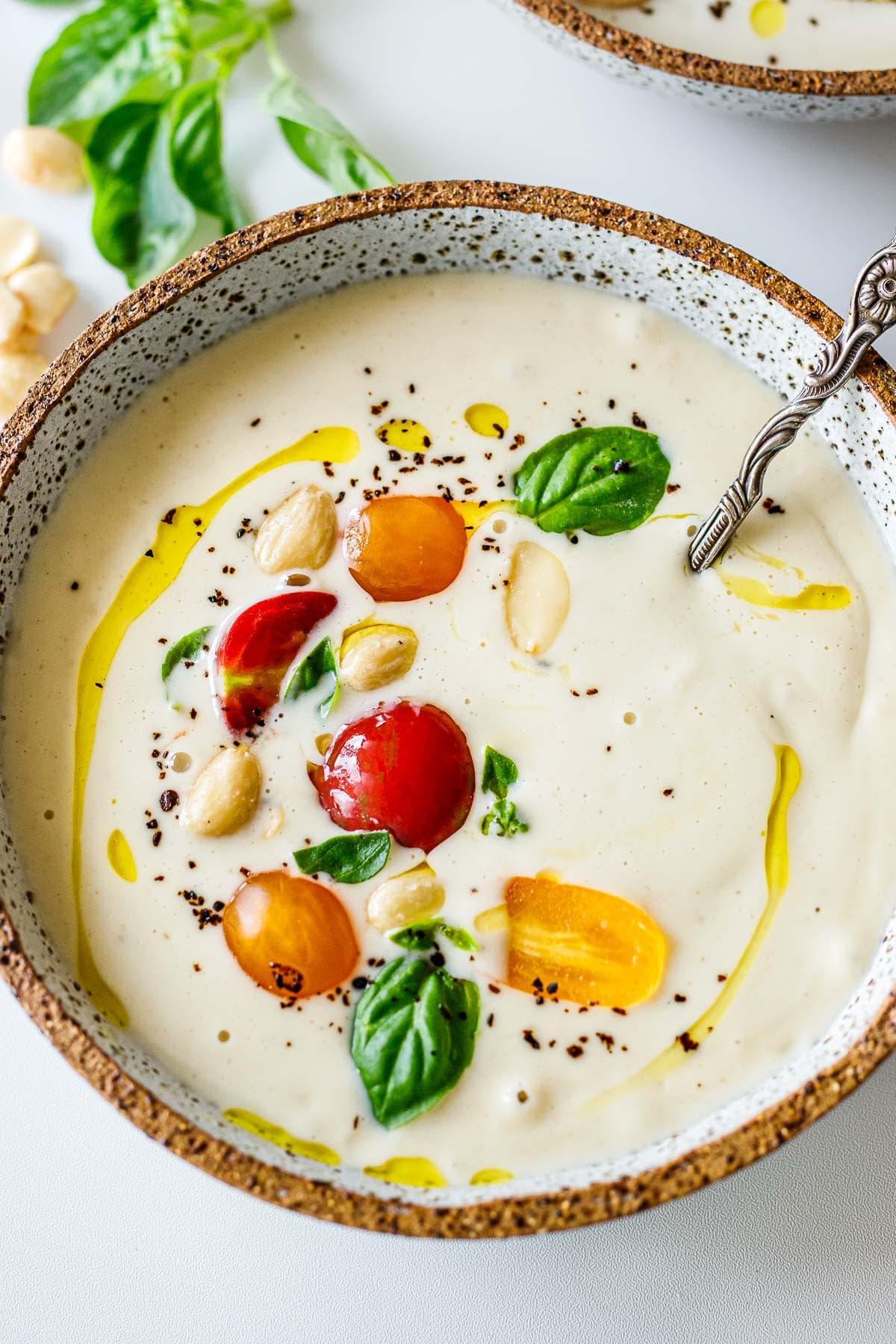soup bowl and spoon with ajo blanco topped with grape tomatoes, marcona almonds, fresh basil, black pepper, and olive oil. 