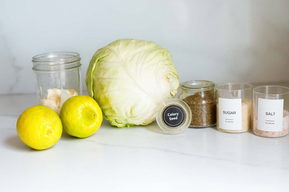 ingredients laid out for creamy coleslaw- mayonnaise, lemons, green cabbage, celery seed, sugar, and salt. 
