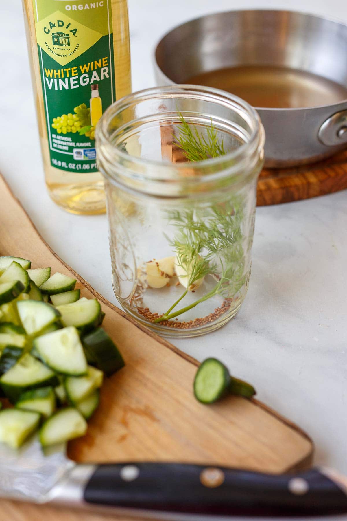 jar with dill, garlic, and mustard seeds next to cutting board with chopped cucumber.